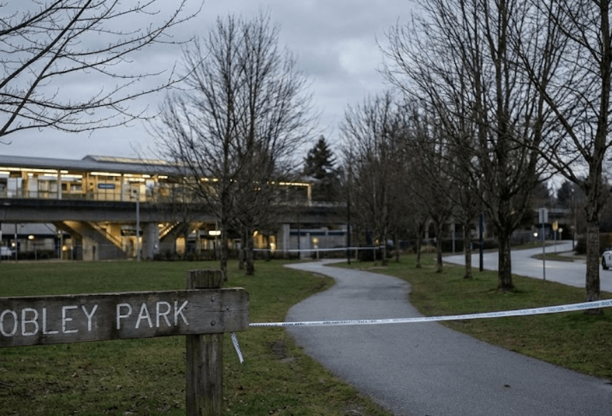 A wide-angle landscape photograph of Lobley Park in Burnaby at dusk, showing police tape blocking a path, a police vehicle with flashing lights, a "POLICE WARNING: STAY VIGILANT" sign, and a SkyTrain station in the background under an overcast sky.
